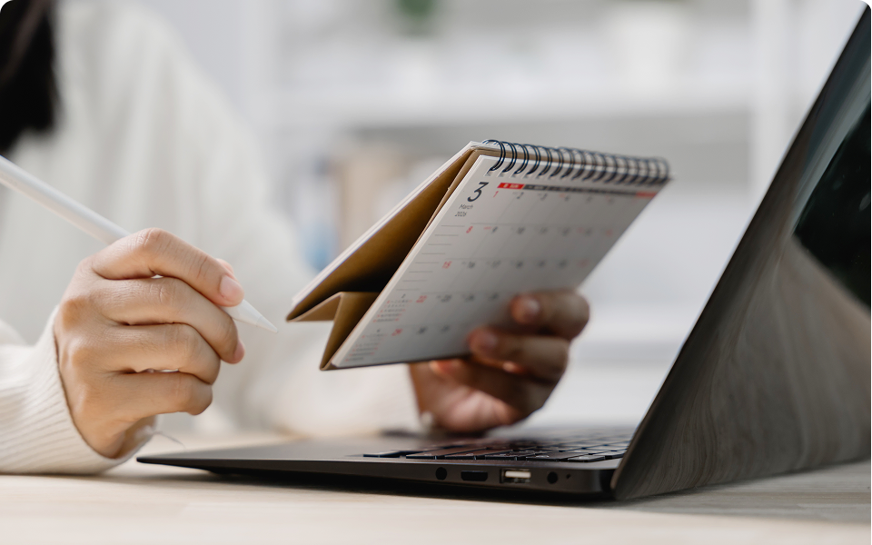 Hand holding a desk calendar in front of a laptop while marking dates with a stylus