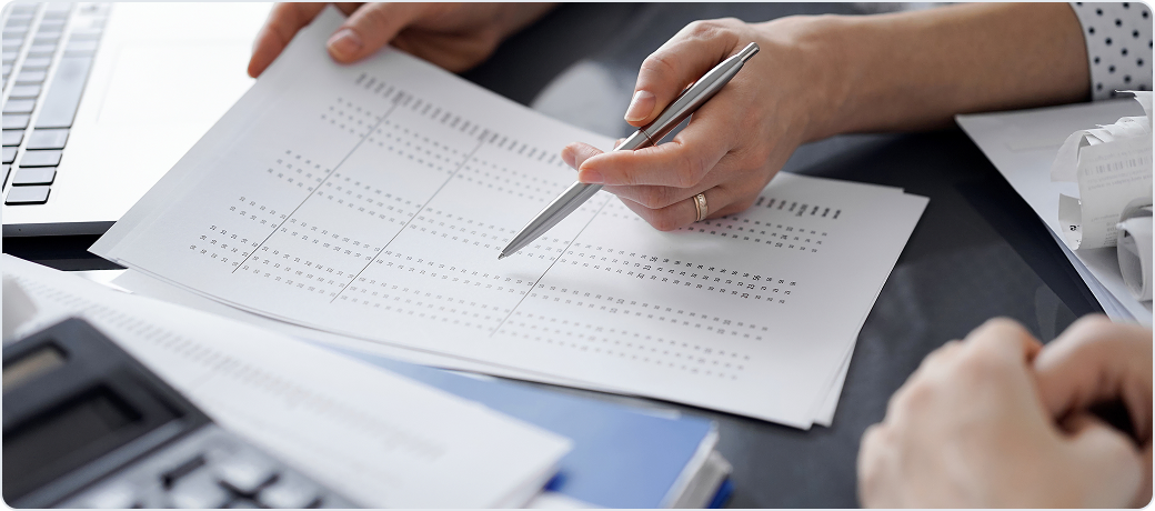 Pen tracing rows of figures on a printed spreadsheet next to a calculator