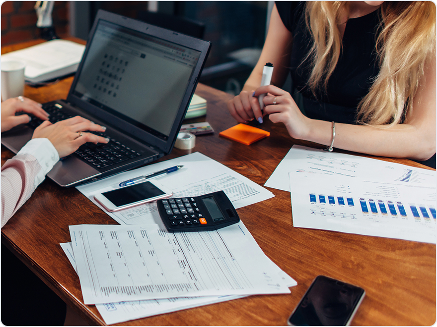 Two colleagues reviewing printed financial reports with a calculator and a laptop at a wooden desk