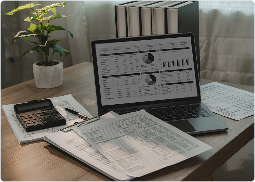 Laptop showing a financial dashboard beside a calculator, printed spreadsheets and a row of binders