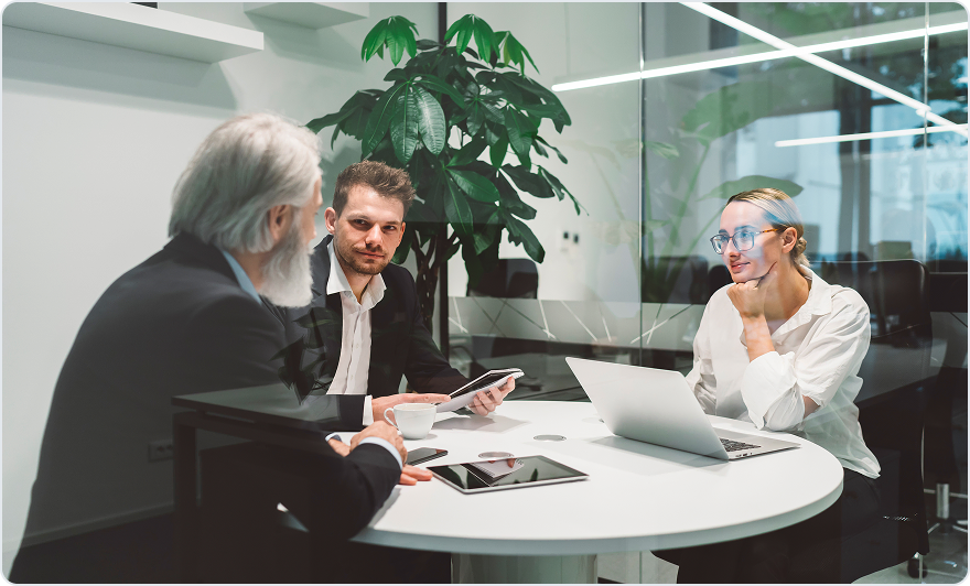 Three people meeting at a round table with a laptop and tablet